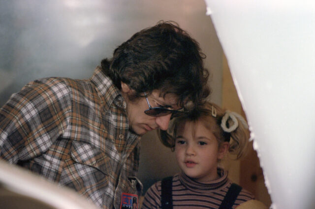 Steven Spielberg leaning into a young Drew Barrymore as she says something to him.