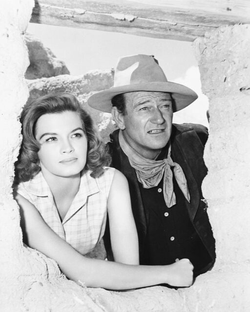 Angie Dickinson and John Wayne peak through a whole in a wall together.