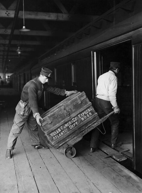 Two workers loading a wooden Wells Fargo crate onto a train. 