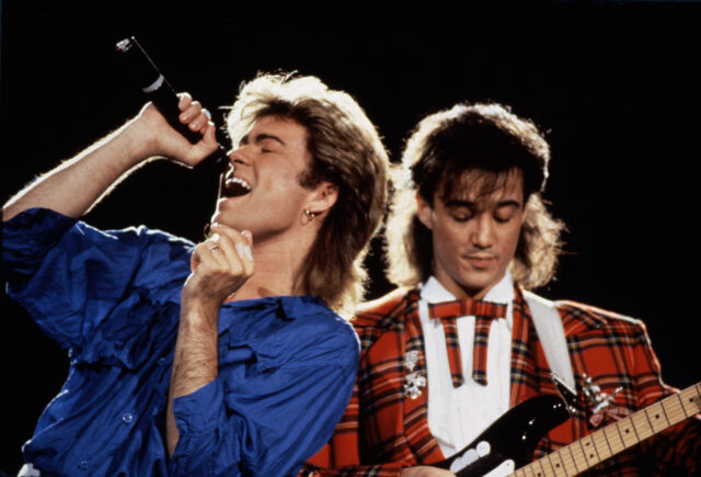 George Michael singing into a microphone while Andrew Ridgeley plays guitar.