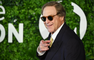 John Goodman poses in sunglasses and a dark suit in front of a bright green background