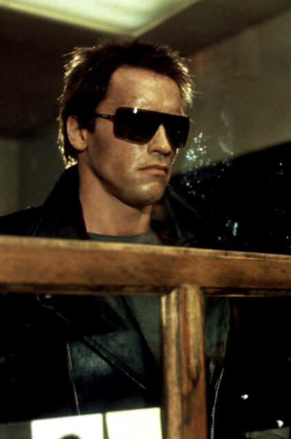 Arnold Schwarzenegger as The Terminator, wearing sunglasses and standing behind a glass window.