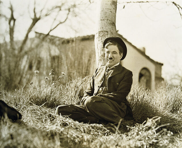 Charlie Chaplin leans against a tree outdoors