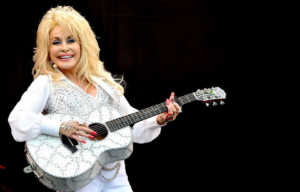 Dolly Parton performing on stage in a white outfit with sequins and matching guitar.