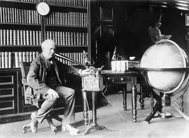 Thomas Edison sits in front of a large bookshelf as he inspects one of his inventions