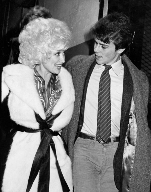 Dolly Parton in a fur jacket walks with David Parton in a jacket and tie, covered with a pea coat. 