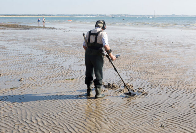Man with a metal detector walks along a beach. 