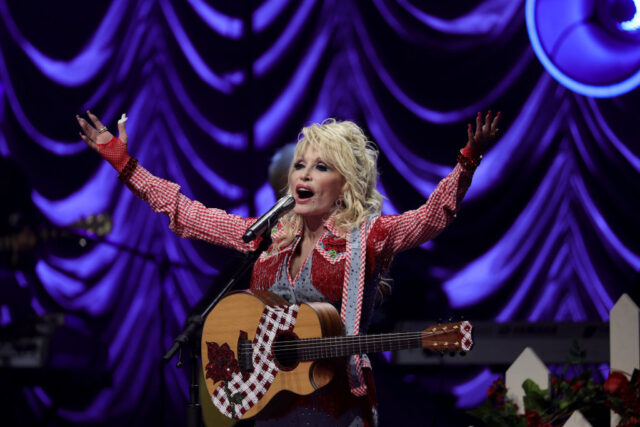 Dolly Parton singing into a microphone with a guitar while wearing a red checked western shirt. 