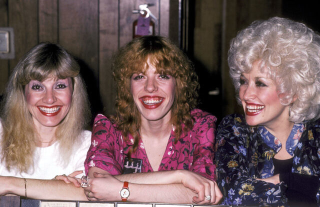 Stella in a white shirt, Freida in a pink floral shirt, and Dolly Parton in a blue floral shirt, smile while standing in a row.