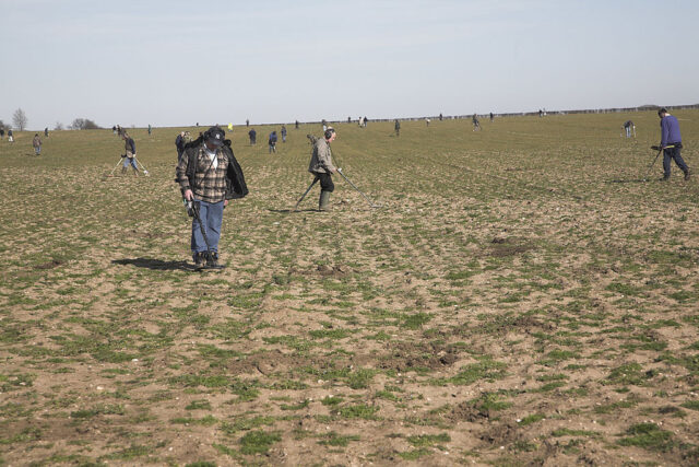 Group of men walk through a field with metal detectors.