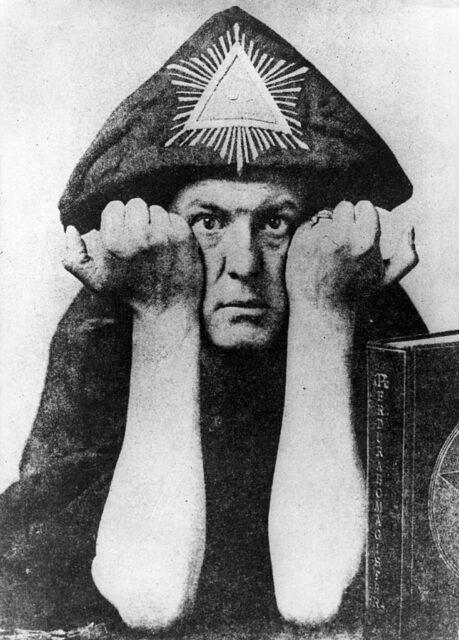 Aleister Crowley in a robe and hat with triangle on it, holding up his forearms to the camera.