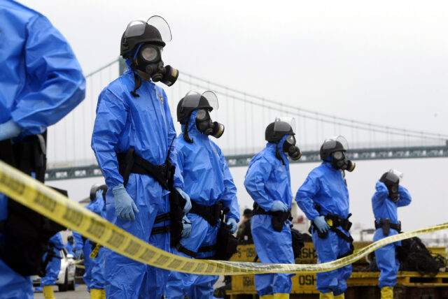 Several people standing in contamination suits behind a caution tape.