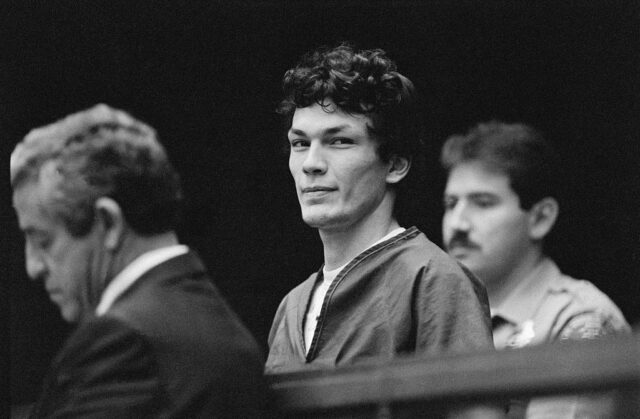 Richard Ramirez in a prison jumpsuit standing beside men in suits.