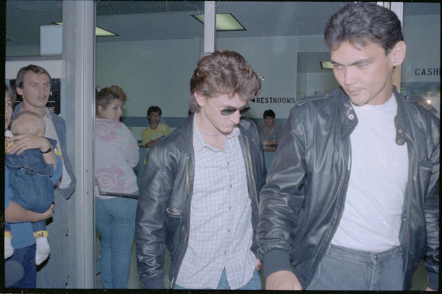 Sean Penn in a leather jacket, light shirt, and jeans, walks behind a friend out of a building.