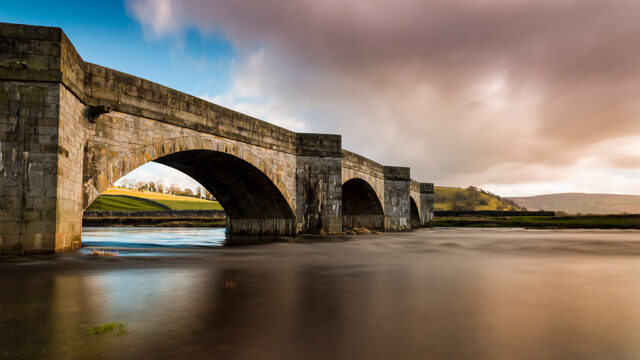 Large stone Burnsall Bridge extending over the River Wharfe in Yorkshire with a slightly pink, cloudy sky.