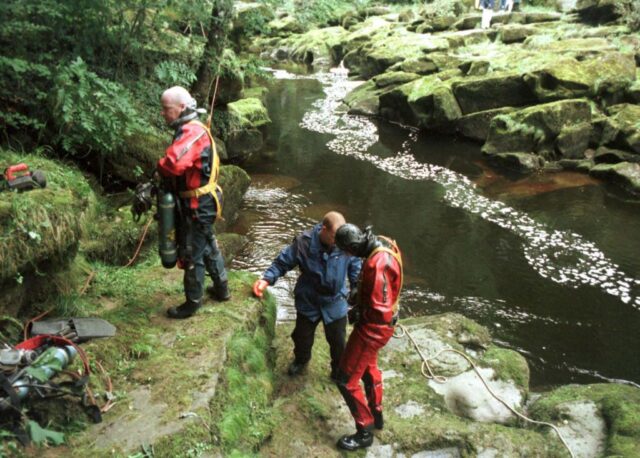 Group of divers in wetsuits stand on the bank of a river. 