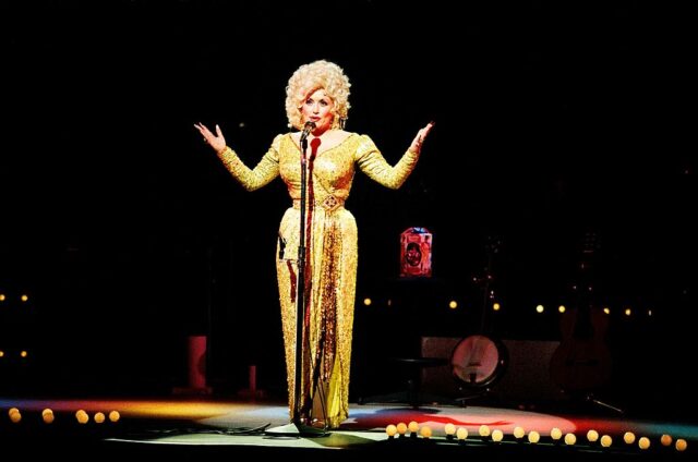 Dolly Parton in stage behind a microphone wearing a golden dress holding her arms up.