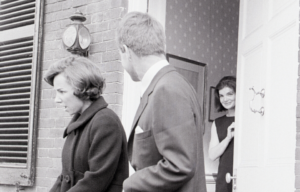 Robert F. Kennedy looks back at Jackie Kennedy smiling at him in a doorway, Ethel Kennedy walking beside him.