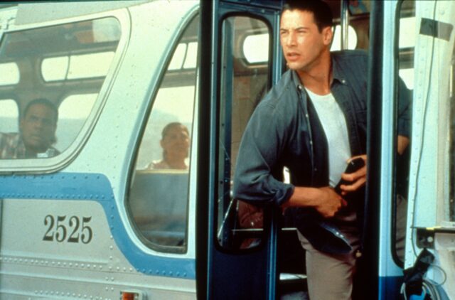 Keanu Reeves as Jack Traven, wearing a denim shirt and brown pants as he leans out an open bus door.