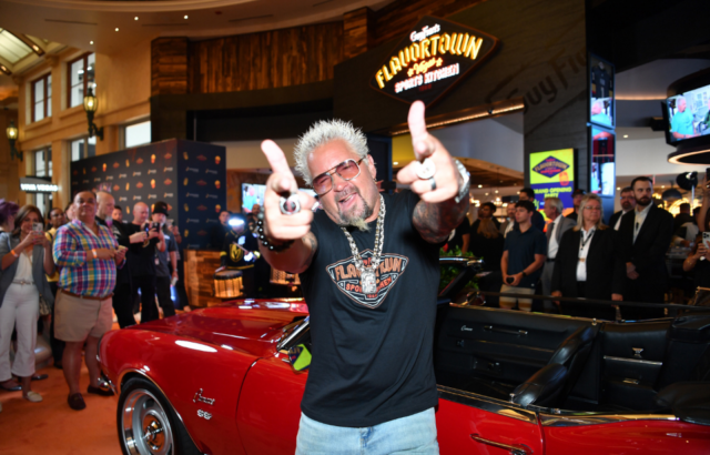 Guy Fieri standing in front of a car, pointing his fingers at the camera
