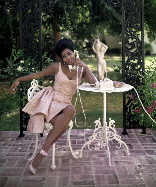 Woman in a pink dress sitting at an outdoor table talking on a rotary phone.