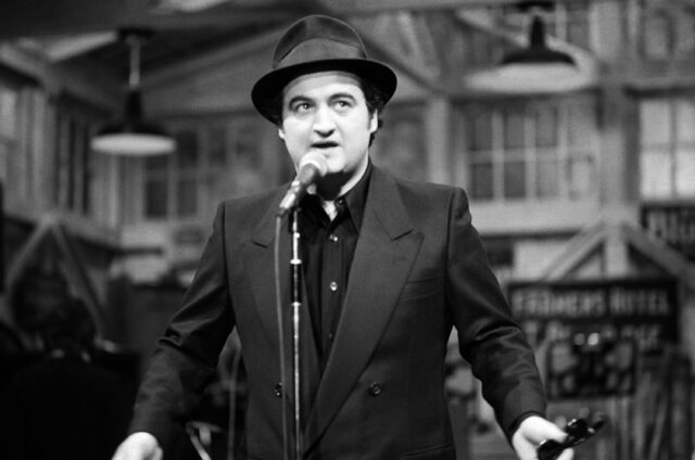 John Belushi standing at a microphone