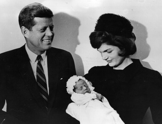 Jackie Kennedy holding a baby, John F. Kennedy standing beside her.