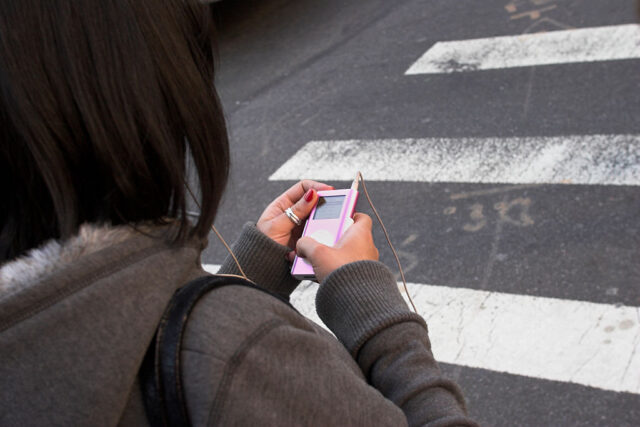 Woman walking across the street while holding her pink iPod in her hands.