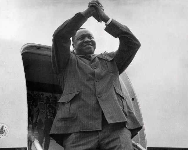 Idi Amin Dada Oumee clasping his hands in front of the entryway to an aircraft