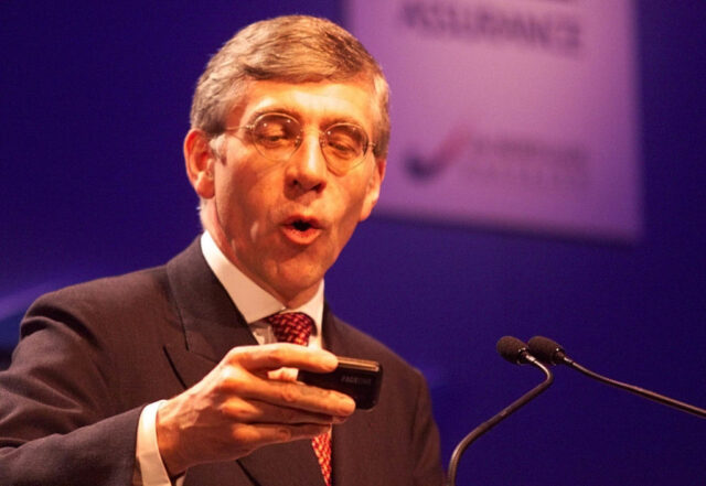 Jack Straw stands behind a microphone while looking at a pager.
