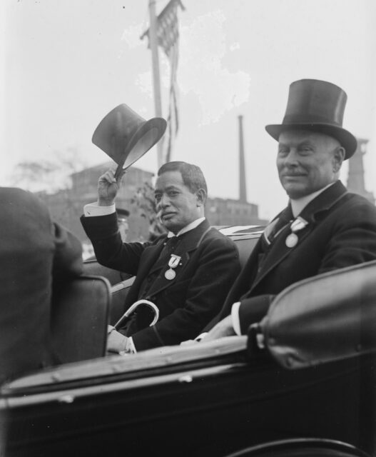 Shirō Ishii tipping his hat while sitting in the back of a vehicle with another man
