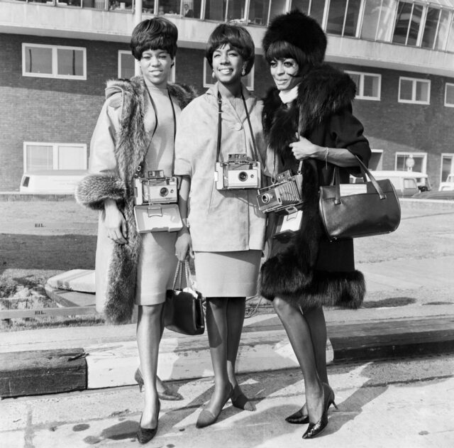 The Supremes posing in fur coats with polaroid cameras hanging around their necks. 