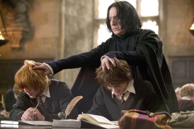 Alan Rickman pushing the heads of Daniel Radcliffe and Rupert Grint toward their desk in a scene from Harry Potter.