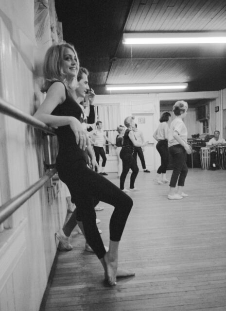 Goldie Hawn in a dance studio, leaning on a barre. Other dancers in the room with her.