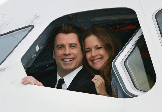John Travolta and Kelly Preston smiling out of the window of an airplane.