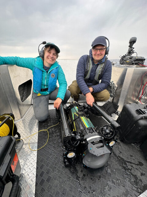 A man and woman crouch on a boat with a remotely operated vehicle between them. 