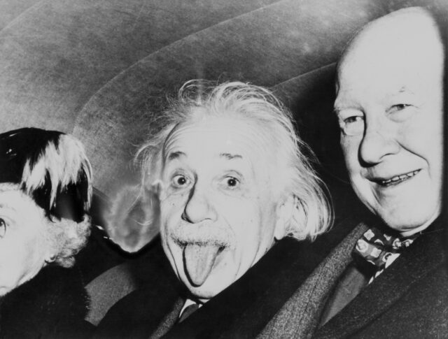 Albert Einstein sitting in a car with two others, sticking his tongue out.