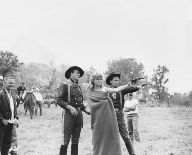 Constance Towers practicing shooting, Johny Wayne, William Holden, and other watch.
