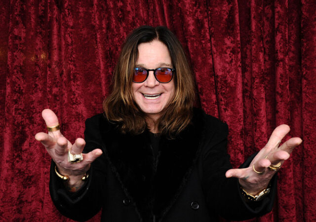 Ozzy Osbourne standing with his arms outstretched in front of a red velvet curtain