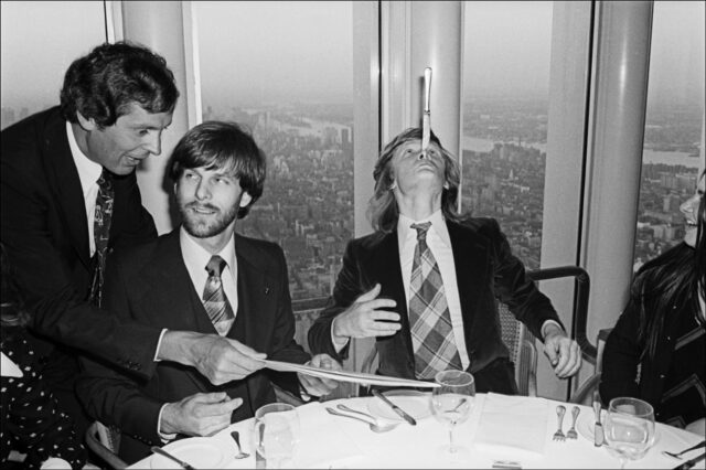 A waiter is handing a menu to George Willig while Philippe Peitit balances a butter knife on his nose.