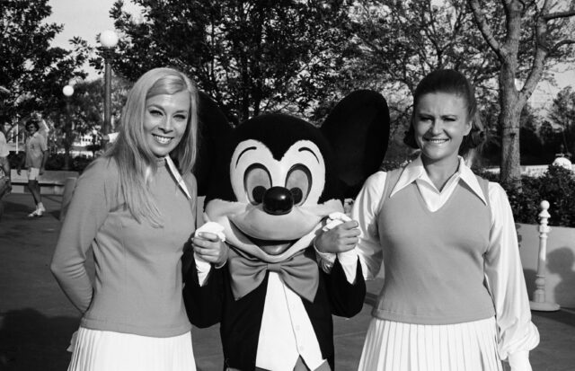 Two women on either side of a Mickey Mouse costume.
