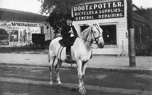 A young boy sitting on a horse, shops in the background.