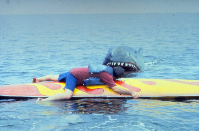 A man laying on a surfboard, a mechanical shark in the background.