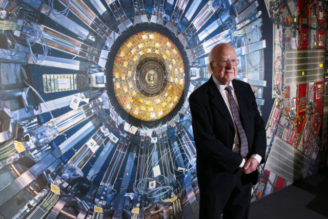 Peter Higgs standing in front of a photograph of a Large Hadron Collider
