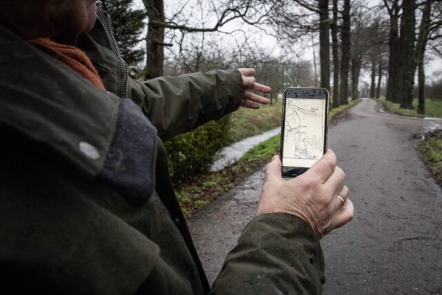 A man holds a cell phone, pointing in the direction in front of him.