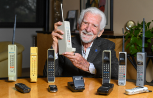 Martin Cooper holds an old cellphone, a table in front of him hold even more old cellphones.