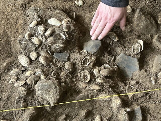 A hand points to pottery pieces buried in dirt.