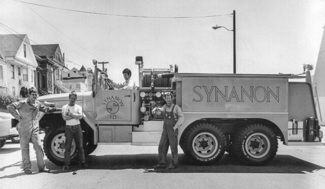 Men stand by a fire truck that has "SYNANON" written on the side. 