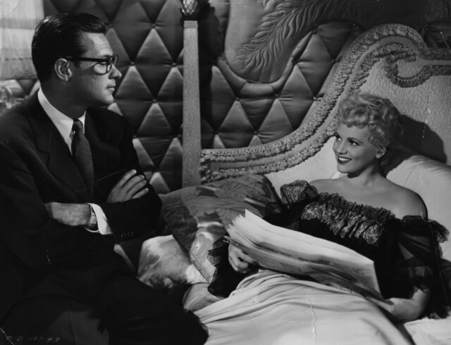 Judy Holliday laying in bed with a man on sitting on it as well.