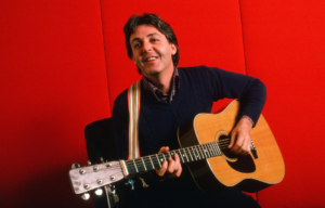 british musician paul mccartney holding guitar smiling against red backdrop